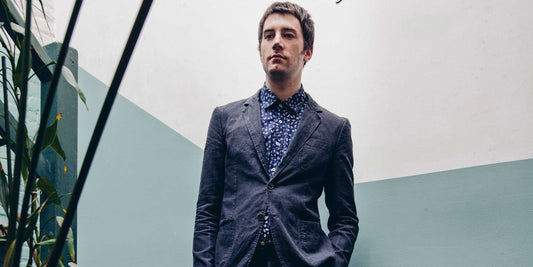 Model stands on a stairway surrounded by plants. He wears a navy Brooklyn Tailors suit and navy patterned dress shirts. 