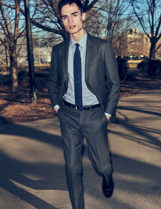 Image of model outside in a park wearing a charcoal suit, light blue shirt, and navy tie. 