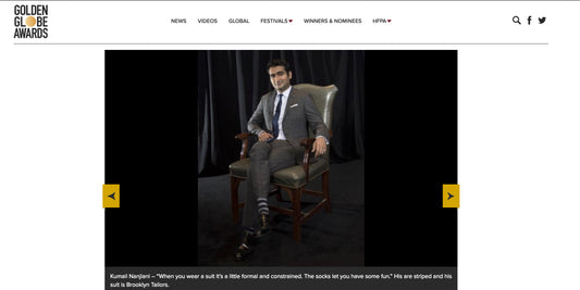Kumail Nanjiani seated in a leather chair wearing a Brooklyn Tailors suit, a white dress shirt, tie, and striped socks. 