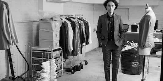 Brooklyn Tailors owner, Daniel Lewis, stands in a basement surrounded by rolling racks, a steamer, and a suit form. He is wearing a dark suit. 