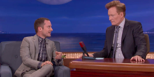 Still from Conan of Elijah Wood wearing a gray suit and blue/gray plaid shirt from Brooklyn Tailors seated next to Conan O'Brien on his late night show. 