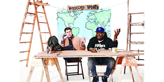 Colin Jost and Michael Che sit at a desk with feet up wearing Brooklyn Tailors 