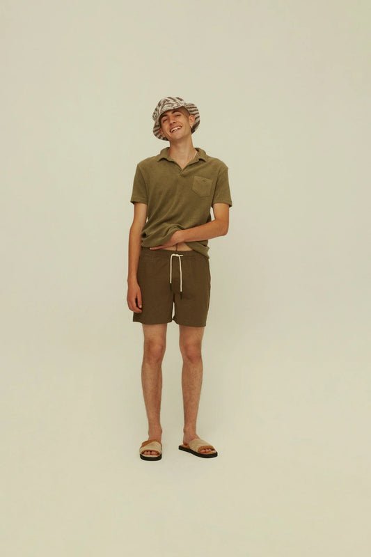 On-body front shot of OAS Army Linen Shorts. Model is wearing an olive terry polo, bucket hat, and slide shoes with the swim trunks. 
