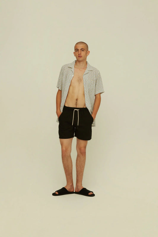 OAS Black Linen Shorts on-body shot. Model is wearing shorts with open camp shirt and slide shoes