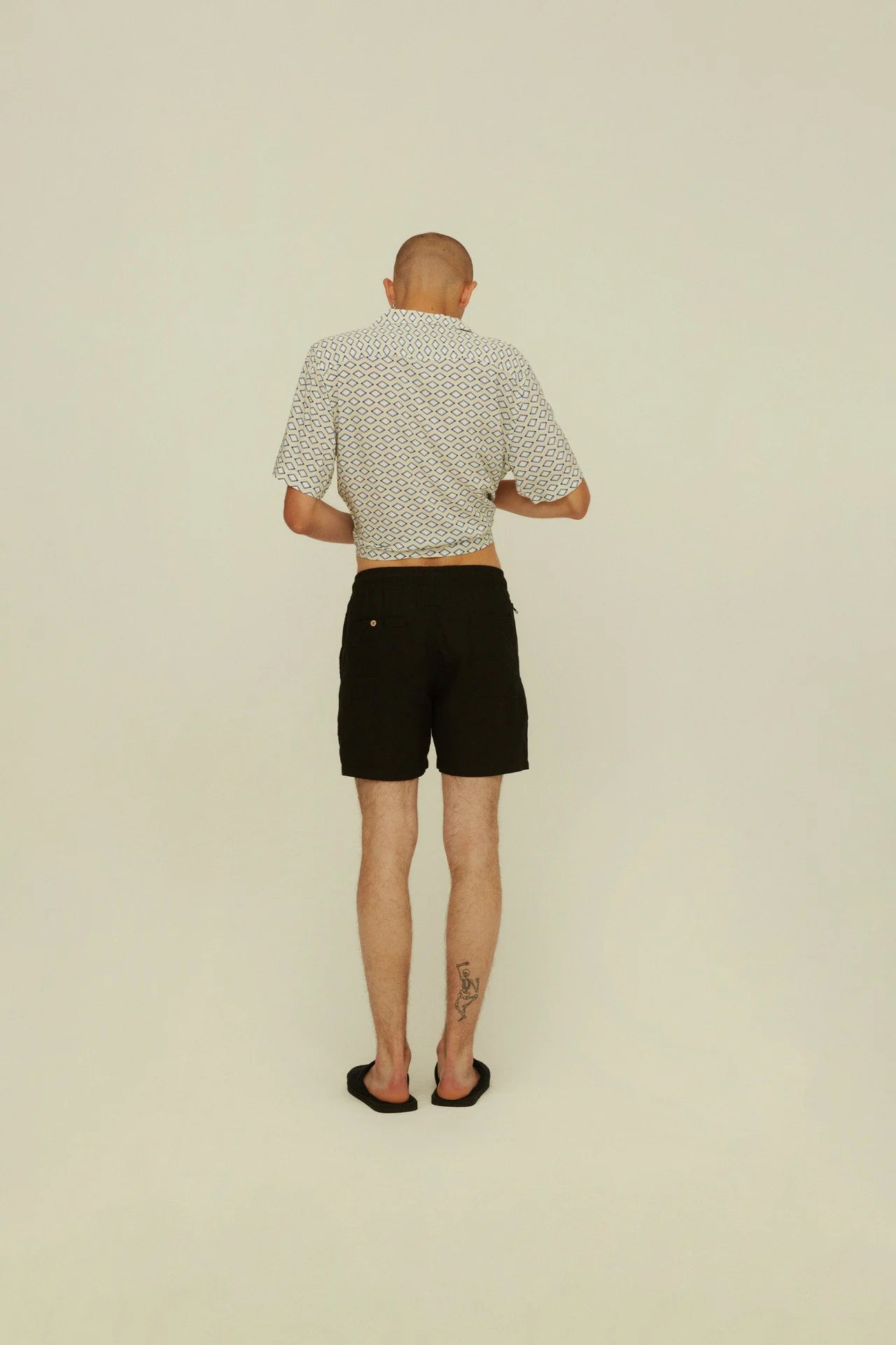 Back on-body shot of Black Linen Shorts. Model is wearing shorts with camp shirt and black slide shoes