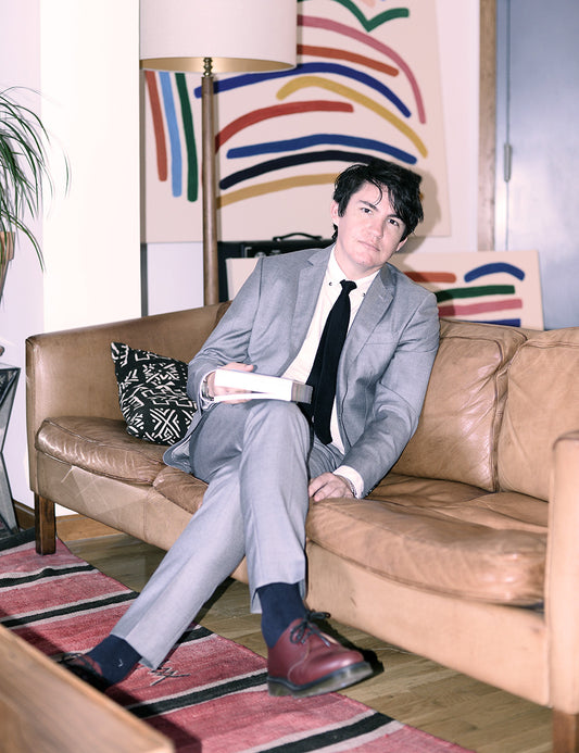 David wears a dove gray suit and sits on a leather couch. 