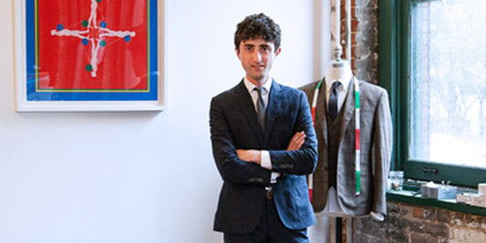 Brooklyn Tailors co-owner, Daniel Lewis, stands in front of a suit form and stacked dress shoes. He wears a navy Brooklyn Tailors suit. 
