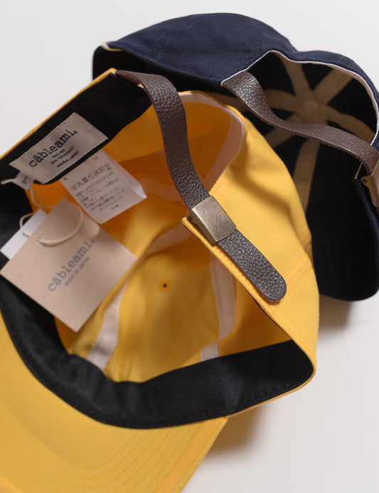 Interior shot of two colors of Cableami Loose Light Chino Baseball Caps showing leather adjuster, labels, and tag