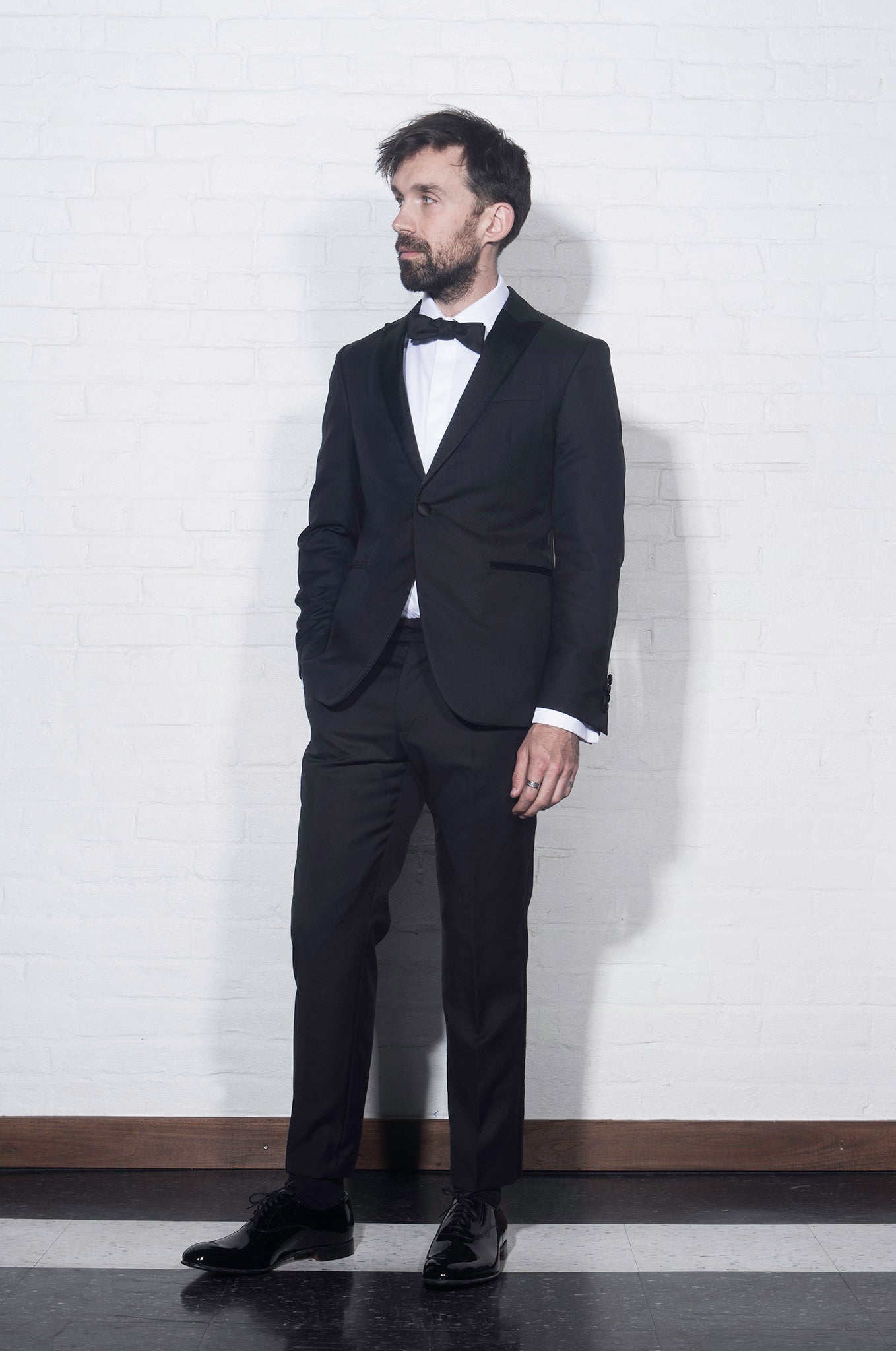 On-body shot of Brooklyn Tailors BKT50 Tuxedo Trouser in Super 110s - Black with Satin Stripe. Model is wearing tuxedo trouser with matching jacket, white tuxedo shirt, and black bowtie. Photo taken from side