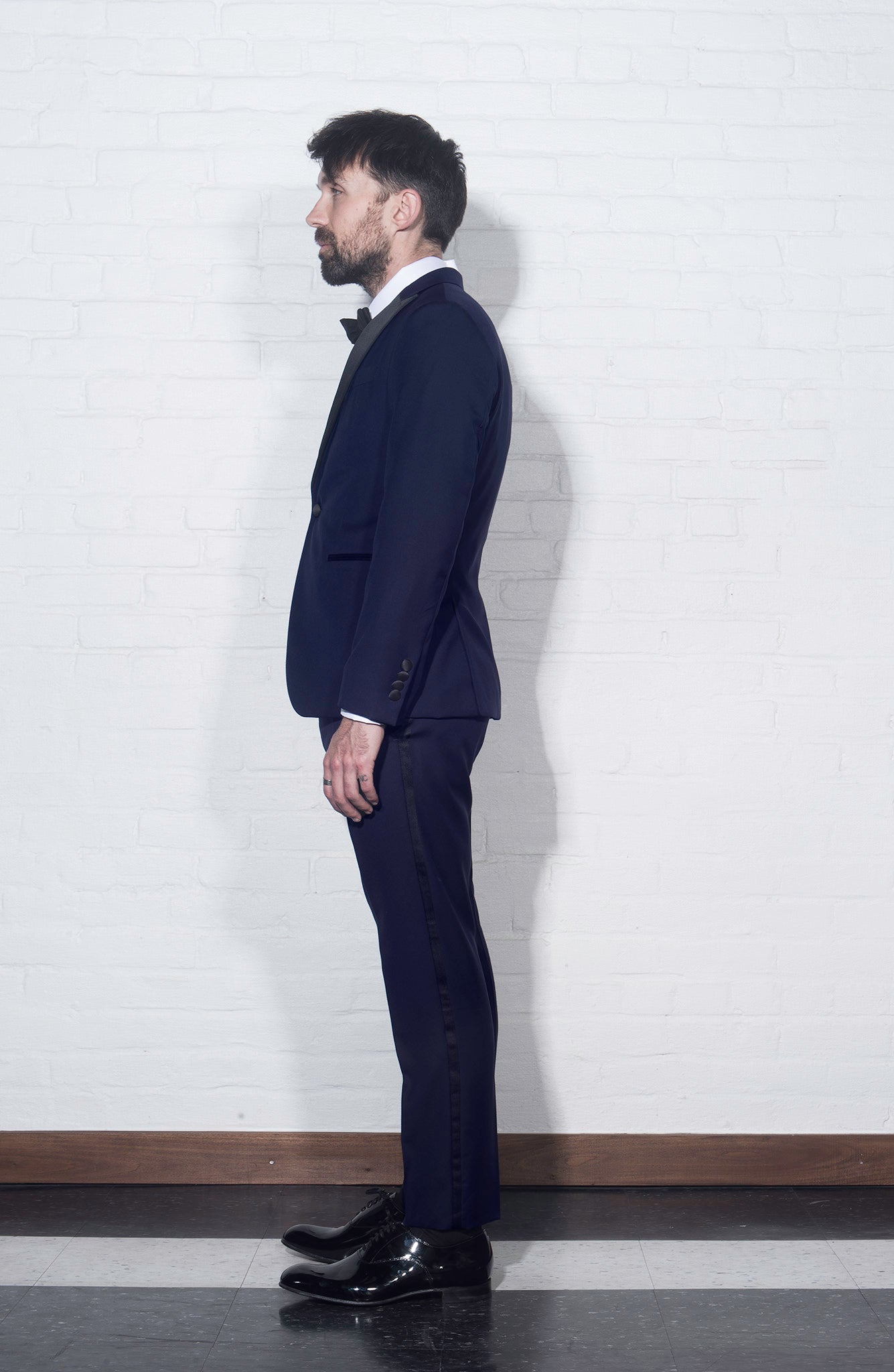On-body shot of BKT50 Tuxedo Trouser in Super 110s - Navy with Satin Stripe from the side. Model is wearing trouser with matching jacket, white tuxedo shirt, and black bowtie