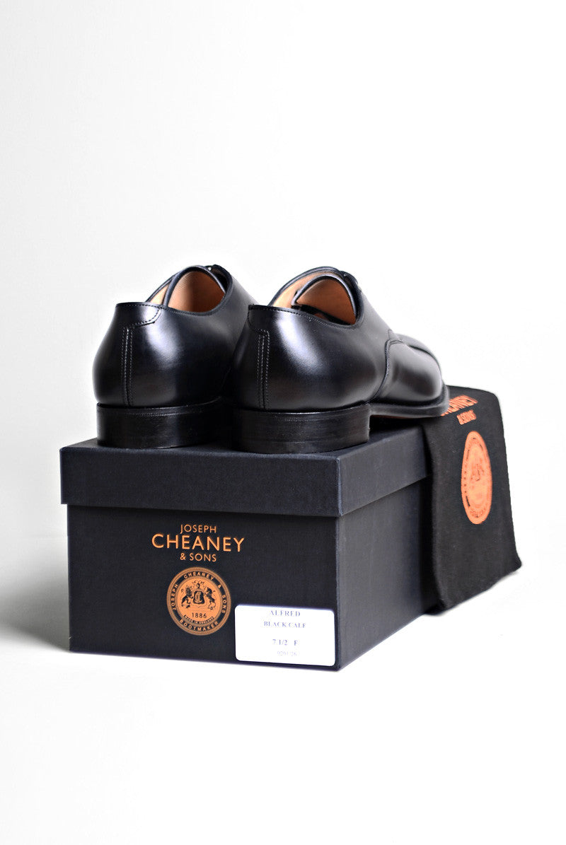 Both Joseph Cheaney Alfred Capped Oxford in Black Calf Leather shoes on top of the box they come in and the dust bag the shoes are sold in. 